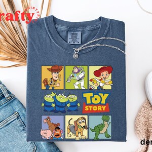 Pu&ograve; includere: Una maglietta blu con una grafica multicolore di personaggi del franchise di film Toy Story. La grafica include Woody, Buzz Lightyear, Jessie e i tre personaggi alieni. Il testo "Toy Story" &egrave; stampato in lettere gialle sotto i personaggi.