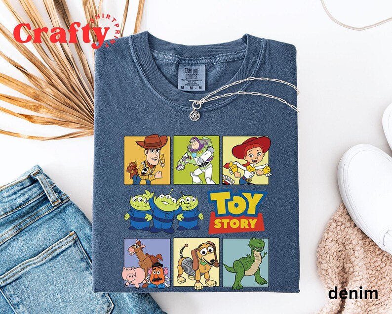 Pu&ograve; includere: Una maglietta blu con una grafica multicolore di personaggi del franchise di film Toy Story. La grafica include Woody, Buzz Lightyear, Jessie e i tre personaggi alieni. Il testo "Toy Story" &egrave; stampato in lettere gialle sotto i personaggi.