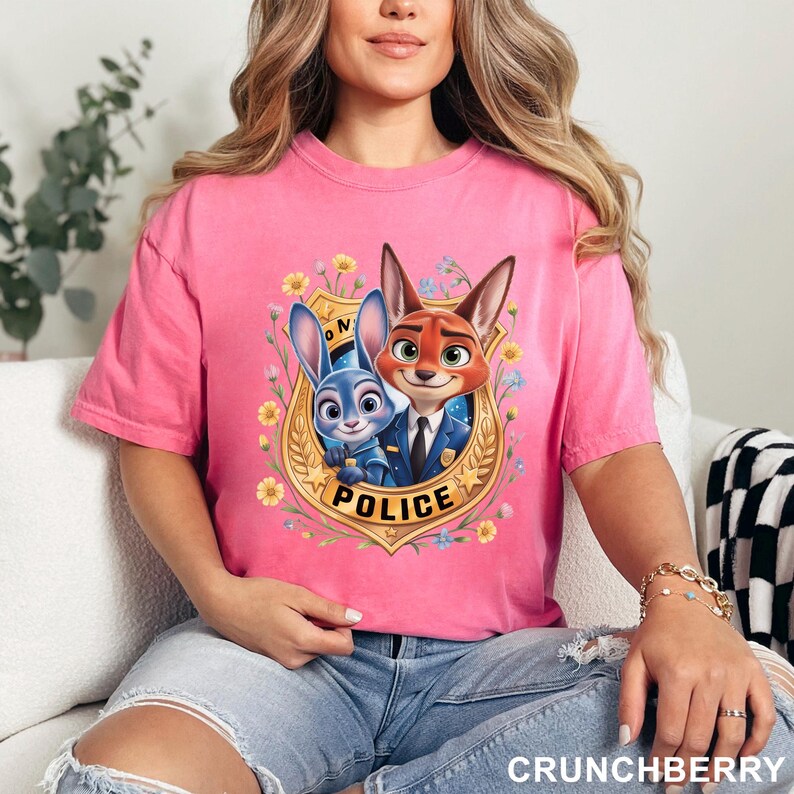 May include: A pink t-shirt featuring a cartoon image of two animal characters from the movie Zootopia, a bunny and a fox, inside a police badge. The word "POLICE" is printed below the characters. The shirt is made of a soft material.