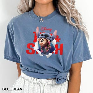 Puede incluir: Una camiseta azul vaquera con un gr&aacute;fico de Stitch de Lilo & Stitch de Disney. El gr&aacute;fico muestra a Stitch rompiendo un agujero con un mono marr&oacute;n. Tambi&eacute;n se ve el texto "Disney" y "Lilo & Stitch".