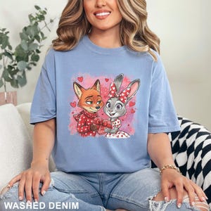 May include: A pale blue t-shirt with a cartoon illustration of a fox and rabbit, surrounded by hearts. The fox is in a red sweater and scarf, and the rabbit has a bow and dress with heart patterns. The text "WASHED DENIM" is at the bottom.