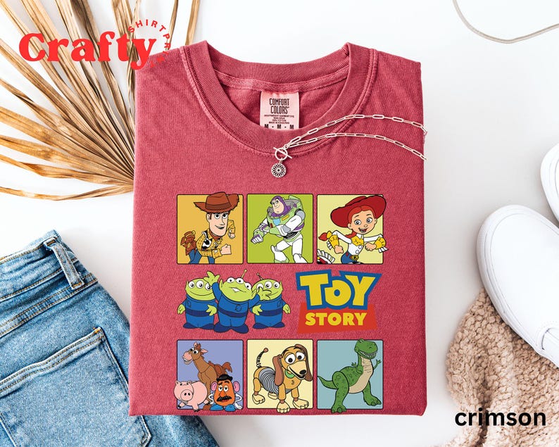 Pu&ograve; includere: Maglietta rossa a tema Toy Story. La maglietta presenta un collage di personaggi del film, tra cui Woody, Buzz Lightyear, Jessie e gli alieni. Il testo "Toy Story" &egrave; stampato in lettere bianche su sfondo rosso.