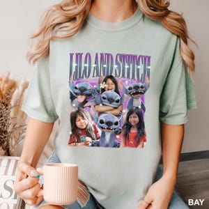 May include: A light green t-shirt featuring the text "Lilo and Stitch" in purple, with a graphic of the characters and children. The shirt has a relaxed fit and short sleeves. The word "BAY" is in the bottom right corner.