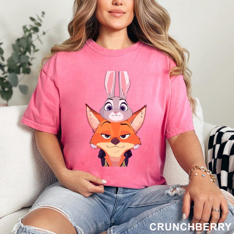 Puede incluir: Camiseta rosa con una ilustraci&oacute;n de dibujos animados de Judy Hopps y Nick Wilde de Zootopia. Los personajes est&aacute;n apilados uno encima del otro. La camiseta tiene mangas cortas y cuello redondo. El texto "CRUNCHBERRY" est&aacute; en la parte inferior derecha.