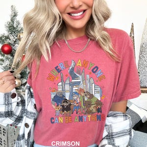May include: Crimson-colored t-shirt featuring a colorful graphic with the text "WHERE ANYONE CAN BE ANYTHING" above a cartoon illustration of various animal characters and city buildings.
