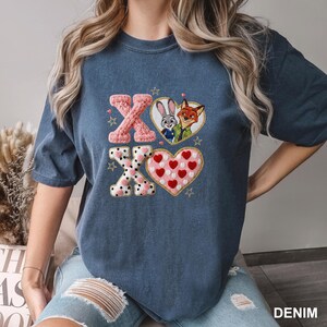 May include: A denim blue t-shirt featuring an embroidered design with the letters "XO" and two hearts. One heart contains the characters Judy Hopps and Nick Wilde. The other heart is filled with red hearts. The word "DENIM" is printed on the bottom right.