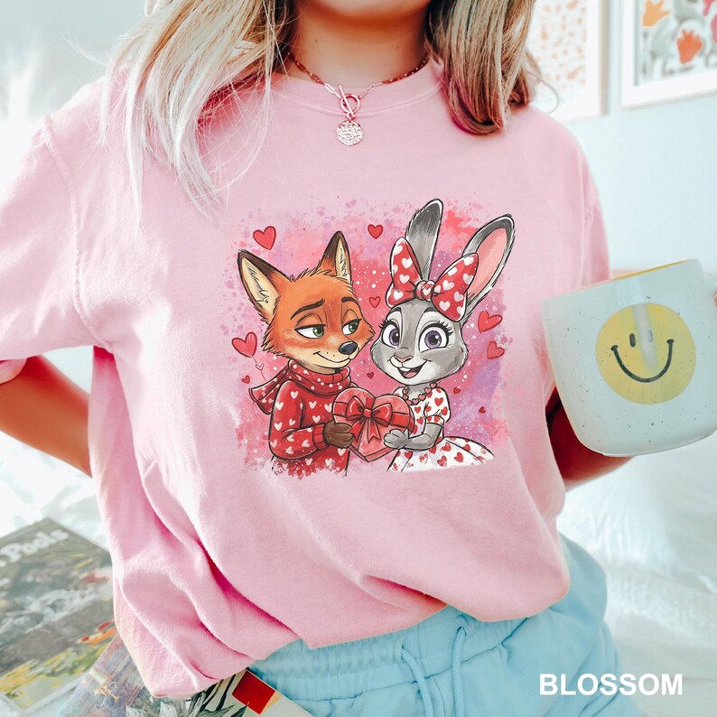 May include: A pale pink t-shirt with a cartoon fox and rabbit design, holding a heart-shaped gift box, with hearts around them. The fox wears a red scarf, and the rabbit has a bow. A white mug with a yellow smiley face is held.