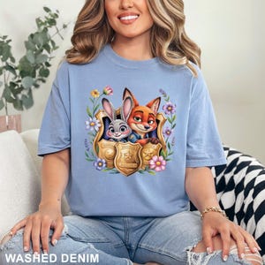 Pu&ograve; includere: T-shirt azzurra con una grafica colorata di Judy Hopps e Nick Wilde di Zootopia, incorniciata da fiori e distintivi della polizia. La maglietta &egrave; realizzata in materiale morbido ed &egrave; etichettata "WASHED DENIM".