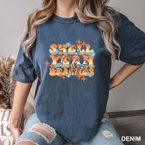 May include: A denim blue t-shirt with the text "Shell Yeah Beaches" in a retro design. The graphic features seashells, palm trees, and stars in orange, yellow, and blue.