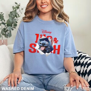 May include: A light blue t-shirt featuring the Disney logo and the words "Lilo & Stitch" in red. The graphic shows Stitch, a blue alien, popping out of a torn paper effect. The shirt is made of a soft material.