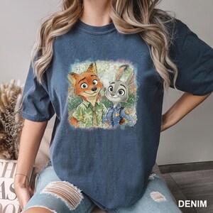 以下が含まれることがあります： デニムブルーのTシャツ。映画「ズートピア」のニック・ワイルドとジュディ・ホップスのカラフルなグラフィックが特徴です。画像には、緑のシャツを着たニックと警察の制服を着たジュディが笑顔で描かれています。「DENIM」の文字が右下に印刷されています。