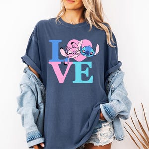 May include: Navy blue t-shirt featuring the word "LOVE" in blue and pink block letters. The "O" is a pink heart with cartoon characters Angel and Stitch. The shirt has a relaxed, oversized fit.