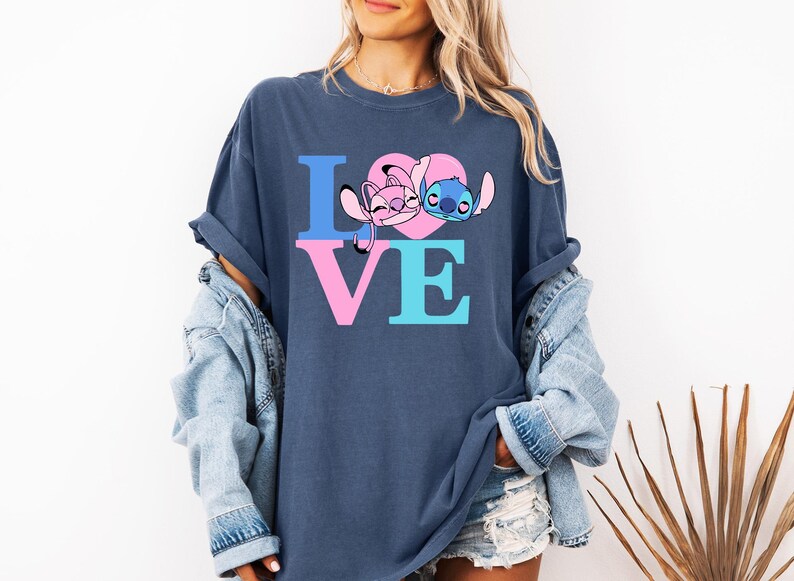 May include: Navy blue t-shirt featuring the word "LOVE" in blue and pink block letters. The "O" is a pink heart with cartoon characters Angel and Stitch. The shirt has a relaxed, oversized fit.
