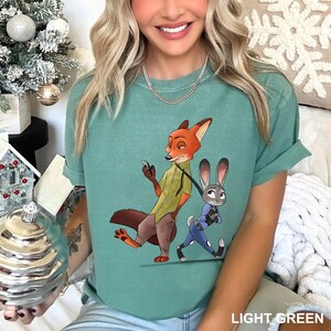 Może przedstawiać: Jasnozielona koszulka z kresk&oacute;wkową ilustracją lisa i kr&oacute;lika. Lis ma na sobie zieloną koszulę i brązowe spodnie, a kr&oacute;lik niebieski mundur. Tekst "LIGHT GREEN" znajduje się na dole.