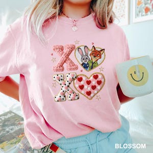 May include: A light pink sweatshirt featuring embroidered "XO" letters and a heart-shaped design with red hearts. The sweatshirt also has a cartoon image of Judy Hopps and Nick Wilde. A mug with a yellow smiley face is held in the image.