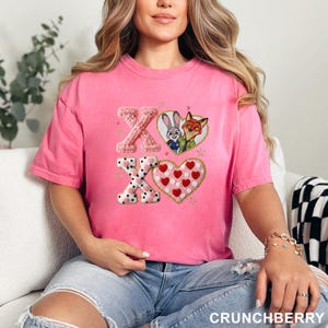 May include: A pink t-shirt featuring embroidered designs. The design includes the letters "XO", a heart with red hearts, and a heart-shaped design with black polka dots. The shirt also features the characters Judy Hopps and Nick Wilde.