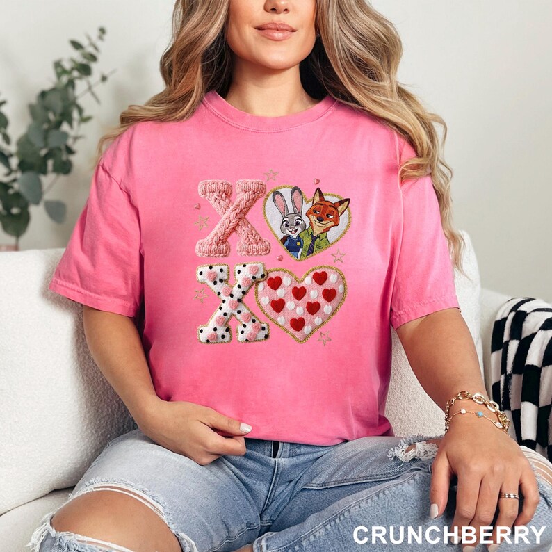 May include: A pink t-shirt featuring embroidered designs. The design includes the letters "XO", a heart with red hearts, and a heart-shaped design with black polka dots. The shirt also features the characters Judy Hopps and Nick Wilde.