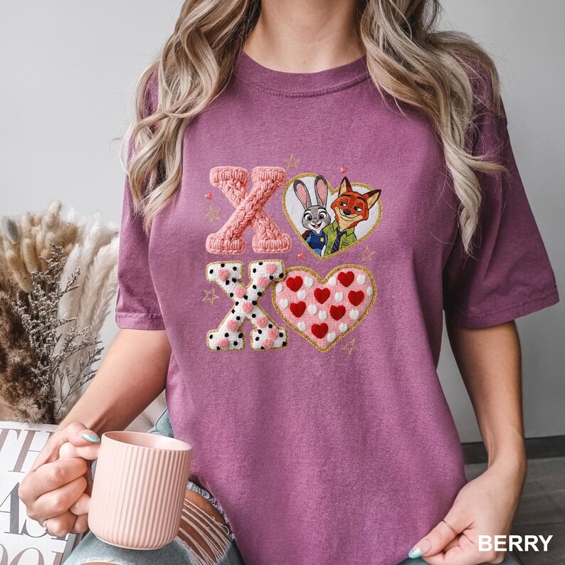 May include: Berry-colored t-shirt featuring an embroidered design with the letters "XO", hearts, and cartoon characters. The "X" letters have a knitted appearance, while the hearts are filled with polka dots and red hearts. The shirt has short sleeves.