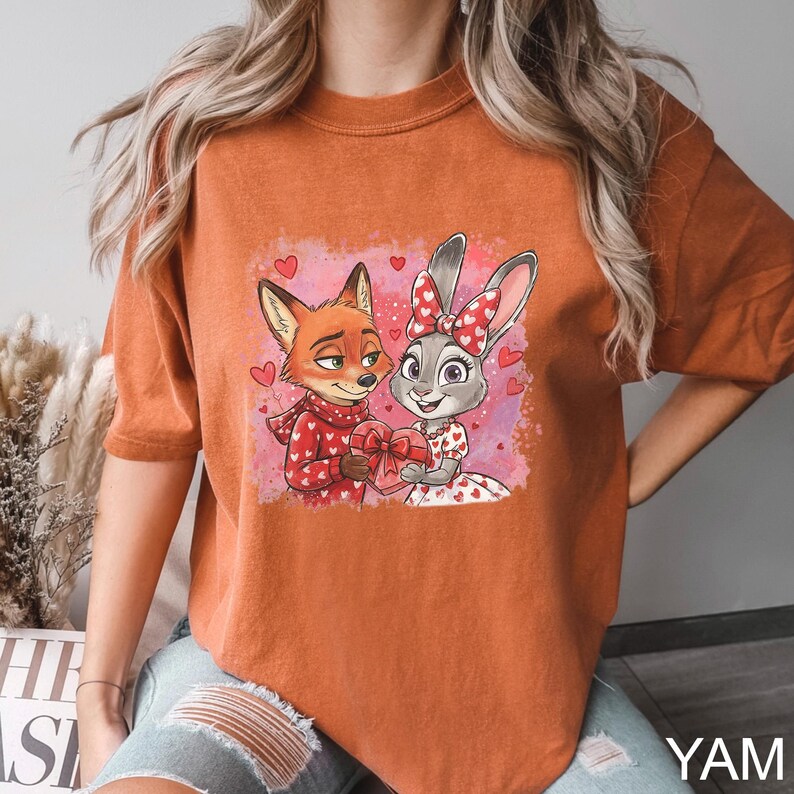 May include: Burnt orange t-shirt with a cartoon fox and bunny sharing a heart-shaped gift. The fox is in a red sweater and scarf, the bunny in a dress and bow with heart patterns. Pink hearts and a pink background complete the design.