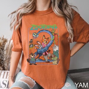 Puede incluir: Camiseta naranja con un dise&ntilde;o gr&aacute;fico colorido de la pel&iacute;cula animada Zootopia. El dise&ntilde;o incluye el t&iacute;tulo de la pel&iacute;cula y varios personajes, como un zorro, un conejo y un perezoso. La camiseta est&aacute; hecha de un material suave.