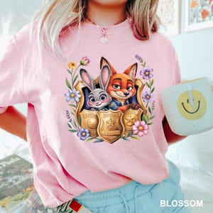 Pu&ograve; includere: T-shirt rosa chiaro con una grafica colorata di Judy Hopps e Nick Wilde di Zootopia all'interno di un distintivo della polizia dorato, circondato da fiori. La maglietta ha anche la scritta "BLOSSOM". Nella foto &egrave; tenuta una tazza bianca con una faccina gialla.