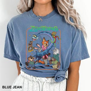 Puede incluir: Camiseta azul vaquero con un gr&aacute;fico colorido de la pel&iacute;cula animada Zootopia. El gr&aacute;fico incluye el t&iacute;tulo de la pel&iacute;cula y varios personajes, como un zorro, un conejo y un perezoso. La camiseta tiene mangas cortas y cuello redondo.