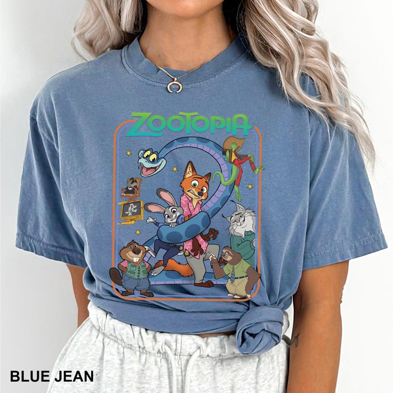 Puede incluir: Camiseta azul vaquero con un gr&aacute;fico colorido de la pel&iacute;cula animada Zootopia. El gr&aacute;fico incluye el t&iacute;tulo de la pel&iacute;cula y varios personajes, como un zorro, un conejo y un perezoso. La camiseta tiene mangas cortas y cuello redondo.
