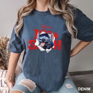 May include: A denim blue t-shirt featuring a graphic of Stitch from Disney's Lilo & Stitch. The design includes the words "Lilo & Stitch" in red, with Stitch appearing to break through the shirt. The Disney logo is at the top.