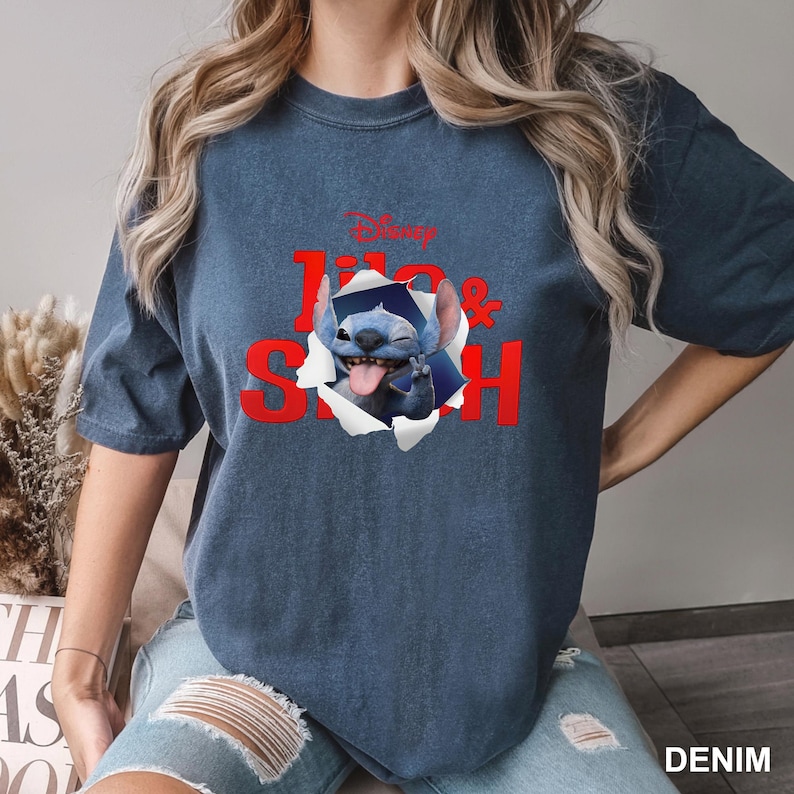 May include: A denim blue t-shirt featuring a graphic of Stitch from Disney's Lilo & Stitch. The design includes the words "Lilo & Stitch" in red, with Stitch appearing to break through the shirt. The Disney logo is at the top.