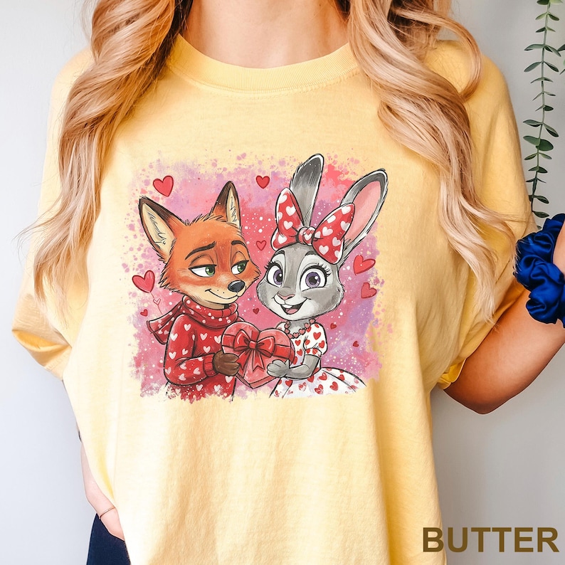 May include: Yellow t-shirt with a cartoon image of a fox and a rabbit, both holding a heart-shaped gift. The fox is in a red sweater with white hearts, and the rabbit wears a dress with heart details. The background is pink with heart accents.