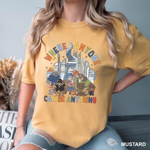 May include: Mustard-colored t-shirt featuring a colorful graphic of various animal characters in front of a cityscape. The text "WHERE ANYONE CAN BE ANYTHING" is displayed above and below the characters in a playful font.