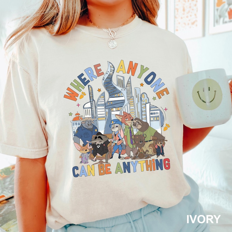 May include: Ivory-colored t-shirt featuring a colorful graphic of cartoon characters and a cityscape. The text "WHERE ANYONE CAN BE ANYTHING" is displayed above the characters. A white mug with a smiley face is held in the image.