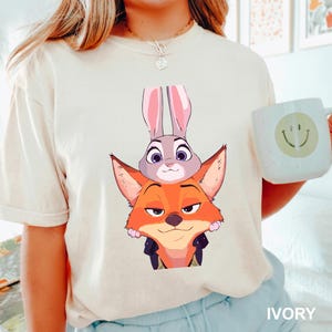 Puede incluir: Camiseta de color marfil con una ilustraci&oacute;n de dibujos animados de Judy Hopps, la conejita, y Nick Wilde, el zorro, de Zootopia. Los personajes est&aacute;n apilados uno encima del otro. La camiseta tiene mangas cortas y cuello redondo. La palabra "IVORY" est&aacute; impresa.