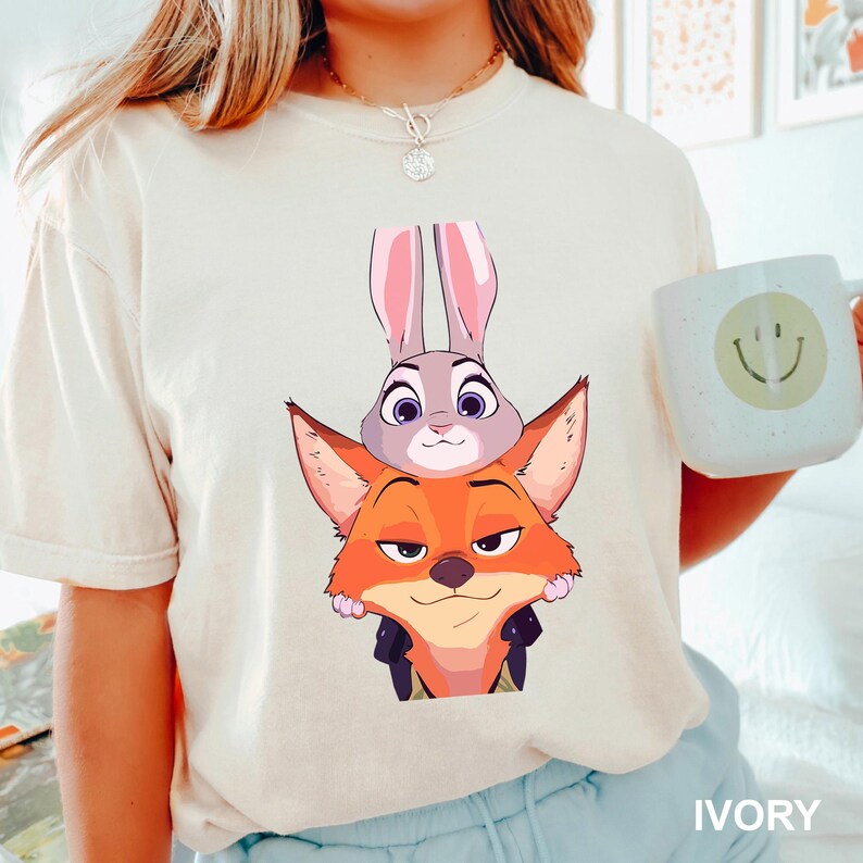 Puede incluir: Camiseta de color marfil con una ilustraci&oacute;n de dibujos animados de Judy Hopps, la conejita, y Nick Wilde, el zorro, de Zootopia. Los personajes est&aacute;n apilados uno encima del otro. La camiseta tiene mangas cortas y cuello redondo. La palabra "IVORY" est&aacute; impresa.