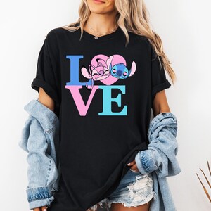 May include: Black t-shirt featuring the word "LOVE" in blue and pink block letters. The "O" is replaced by a pink heart with cartoon characters. The shirt is worn by a person with blonde hair and a denim jacket.