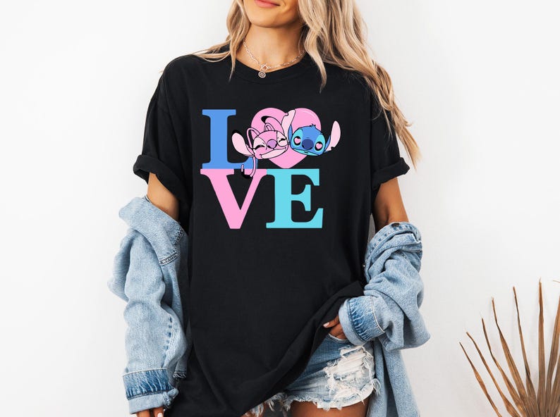 May include: Black t-shirt featuring the word "LOVE" in blue and pink block letters. The "O" is replaced by a pink heart with cartoon characters. The shirt is worn by a person with blonde hair and a denim jacket.
