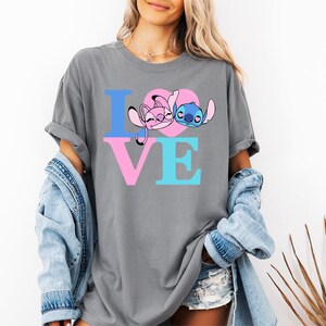 May include: Grey t-shirt featuring the word "LOVE" in blue and pink. The "O" is a pink heart with cartoon characters. This casual t-shirt is part of a clothing collection.