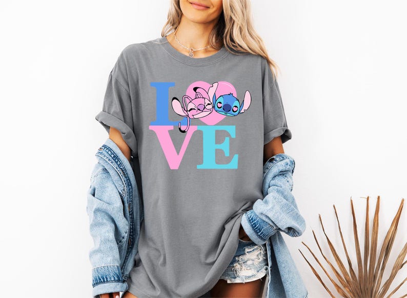 May include: Grey t-shirt featuring the word "LOVE" in blue and pink. The "O" is a pink heart with cartoon characters. This casual t-shirt is part of a clothing collection.