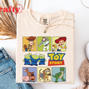 Pu&ograve; includere: Una maglietta beige a tema Toy Story. La maglietta presenta una griglia di nove quadrati con diversi personaggi del film, tra cui Woody, Buzz Lightyear, Jessie e i tre piccoli alieni verdi. Il testo "Toy Story" &egrave; stampato in un font rosso e bianco.