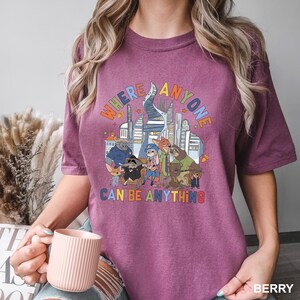 May include: Berry-colored t-shirt featuring a colorful graphic of cartoon characters and a cityscape. The text on the shirt reads "WHERE ANYONE CAN BE ANYTHING" in a rainbow font. The shirt is a short-sleeved crew neck.