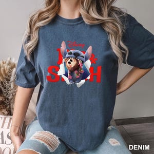 Puede incluir: Camiseta azul denim con un gr&aacute;fico de Stitch y una criatura marr&oacute;n saliendo de un efecto de papel rasgado. La palabra "Disney" est&aacute; encima del gr&aacute;fico, y la palabra "Stitch" es roja.