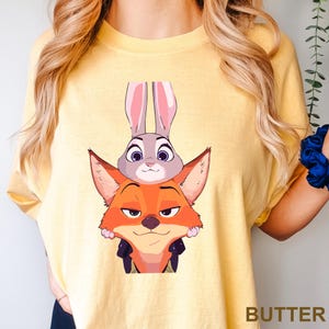 Puede incluir: Camiseta de color mantequilla con una ilustraci&oacute;n de dibujos animados de Judy Hopps, la conejita, y Nick Wilde, el zorro, de Zootr&oacute;polis. Los personajes est&aacute;n apilados. La palabra "BUTTER" est&aacute; impresa en la parte inferior derecha.