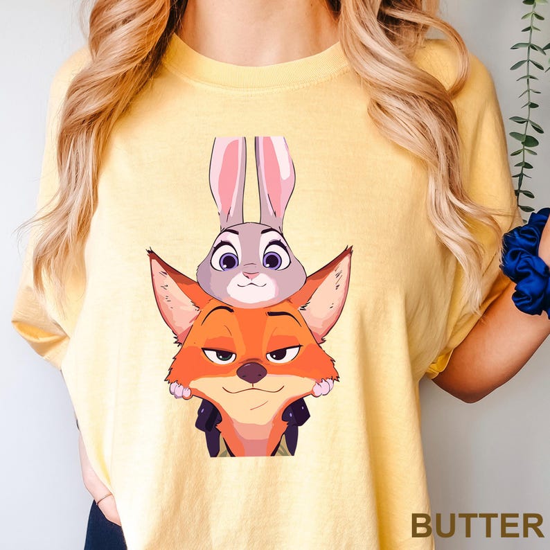 Puede incluir: Camiseta de color mantequilla con una ilustraci&oacute;n de dibujos animados de Judy Hopps, la conejita, y Nick Wilde, el zorro, de Zootr&oacute;polis. Los personajes est&aacute;n apilados. La palabra "BUTTER" est&aacute; impresa en la parte inferior derecha.
