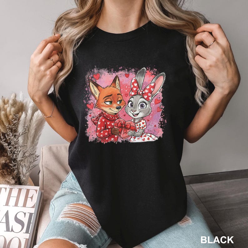May include: Black t-shirt with a cartoon illustration of a fox and a rabbit, both holding a heart-shaped gift. The fox is in a red sweater, and the rabbit wears a dress with red hearts. The background is pink with heart shapes.
