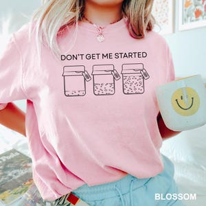 May include: A pale pink sweatshirt featuring the phrase "DON'T GET ME STARTED" above a graphic of three jars with different contents. A white mug with a yellow smiley face is held in the hand.
