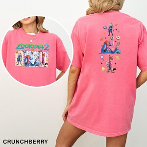 May include: A pink t-shirt featuring the text "ZOOTOPIA 2" with cartoon characters. The back of the shirt has a vertical design with more characters. The shirt is a solid color and has short sleeves. The word "CRUNCHBERRY" is at the bottom.