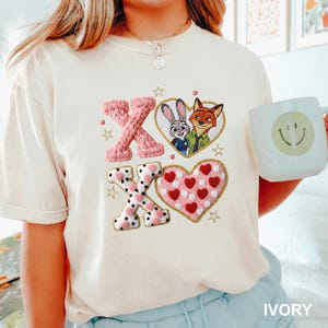 May include: Ivory-colored t-shirt featuring embroidered pink and white X's and a heart with red hearts. The design includes a heart-shaped image of Judy Hopps and Nick Wilde from Zootopia. The shirt also has gold star accents.