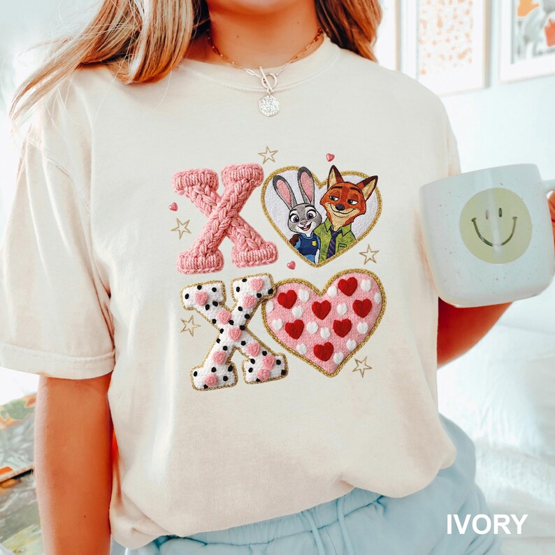 May include: Ivory-colored t-shirt featuring embroidered pink and white X's and a heart with red hearts. The design includes a heart-shaped image of Judy Hopps and Nick Wilde from Zootopia. The shirt also has gold star accents.