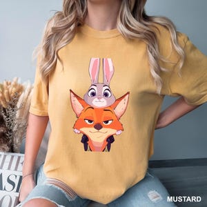 Puede incluir: Camiseta de color mostaza con una ilustraci&oacute;n de dibujos animados de un zorro y un conejo. El zorro es naranja con una expresi&oacute;n astuta, y el conejo es gris con orejas largas. La palabra "MUSTARD" est&aacute; impresa en la parte inferior derecha.