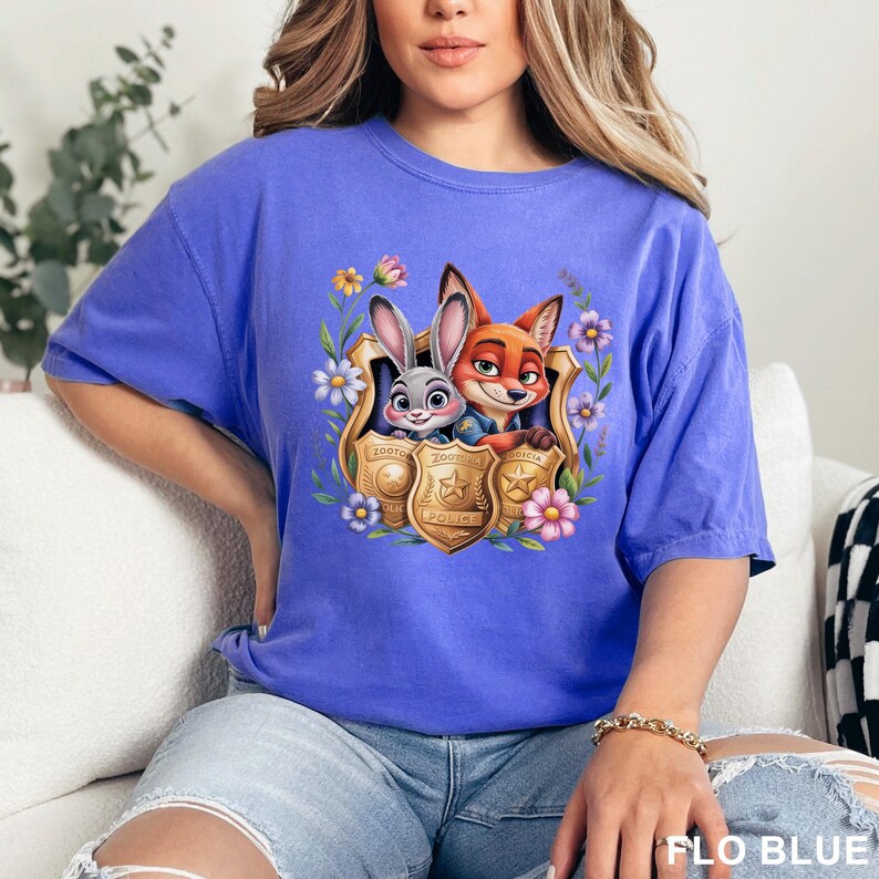 Pu&ograve; includere: T-shirt blu con una grafica di Judy Hopps e Nick Wilde di Zootopia all'interno di un distintivo della polizia, circondata da fiori. Il testo "FLO BLUE" &egrave; in basso a destra.
